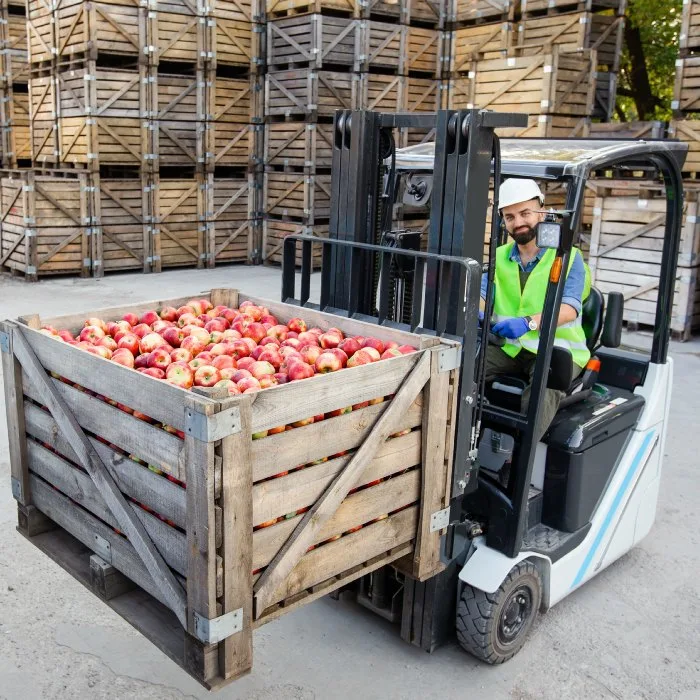 Forklift operator moving a bin of apples past stacked wooden crates