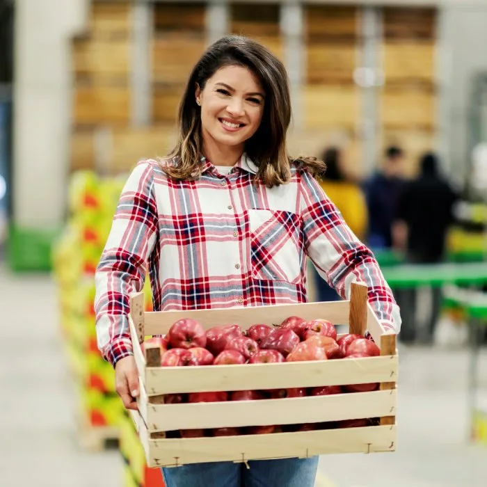 Smiling worker holding a crate of red apples in a packing warehouse